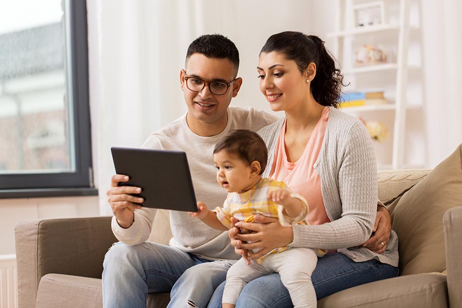 Client Center - Parents Use a Tablet With Their Infant Daughter Perched on Mom's Lap, Sitting on Their Tan Couch in a Bright Living Room