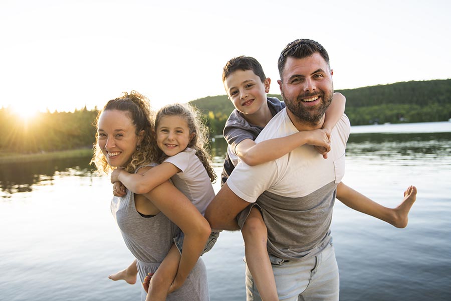 Contact Us - Parents Hold Their Son and Daughter in a Piggyback, Standing on the Shore of a Small Lake at Dusk
